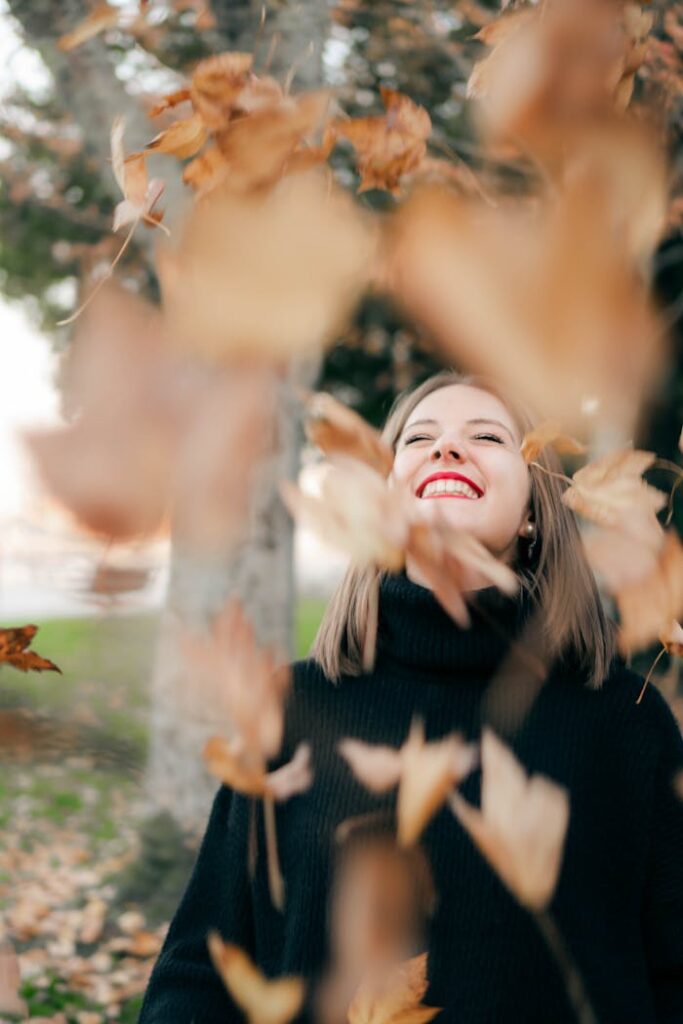 Joyful woman in autumn setting surrounded by falling leaves, embracing the season's beauty.