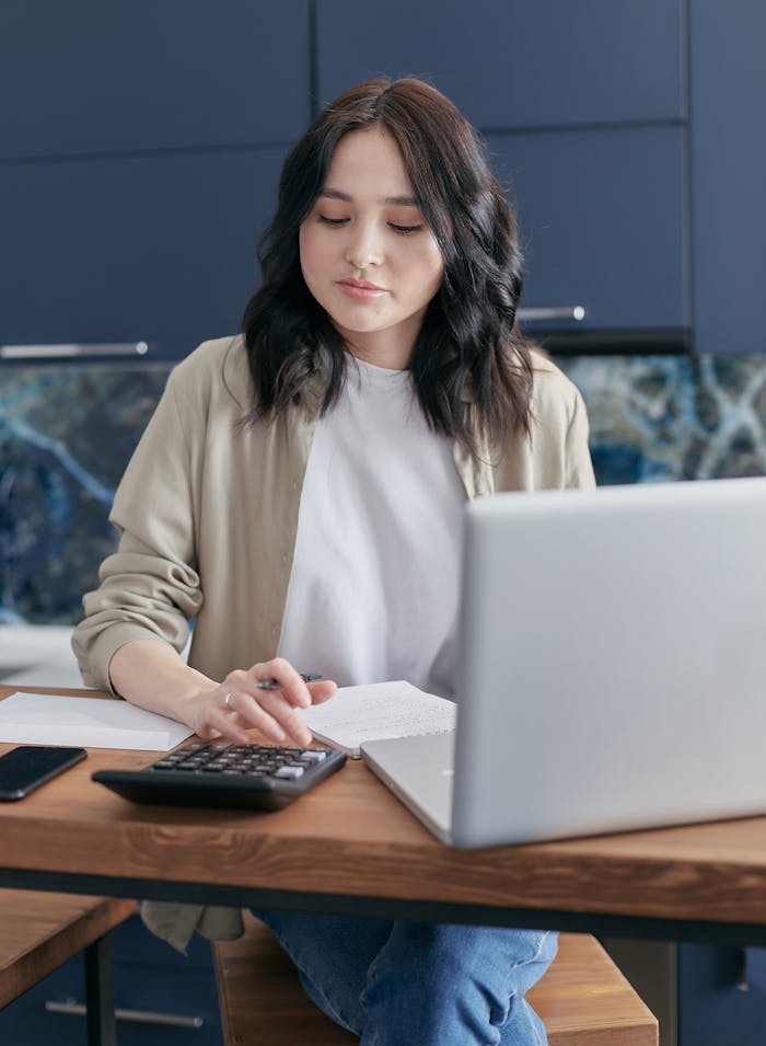 gallery-4 A young woman calculating finances at home using a laptop and calculator, reflecting a focused budgeting process.
