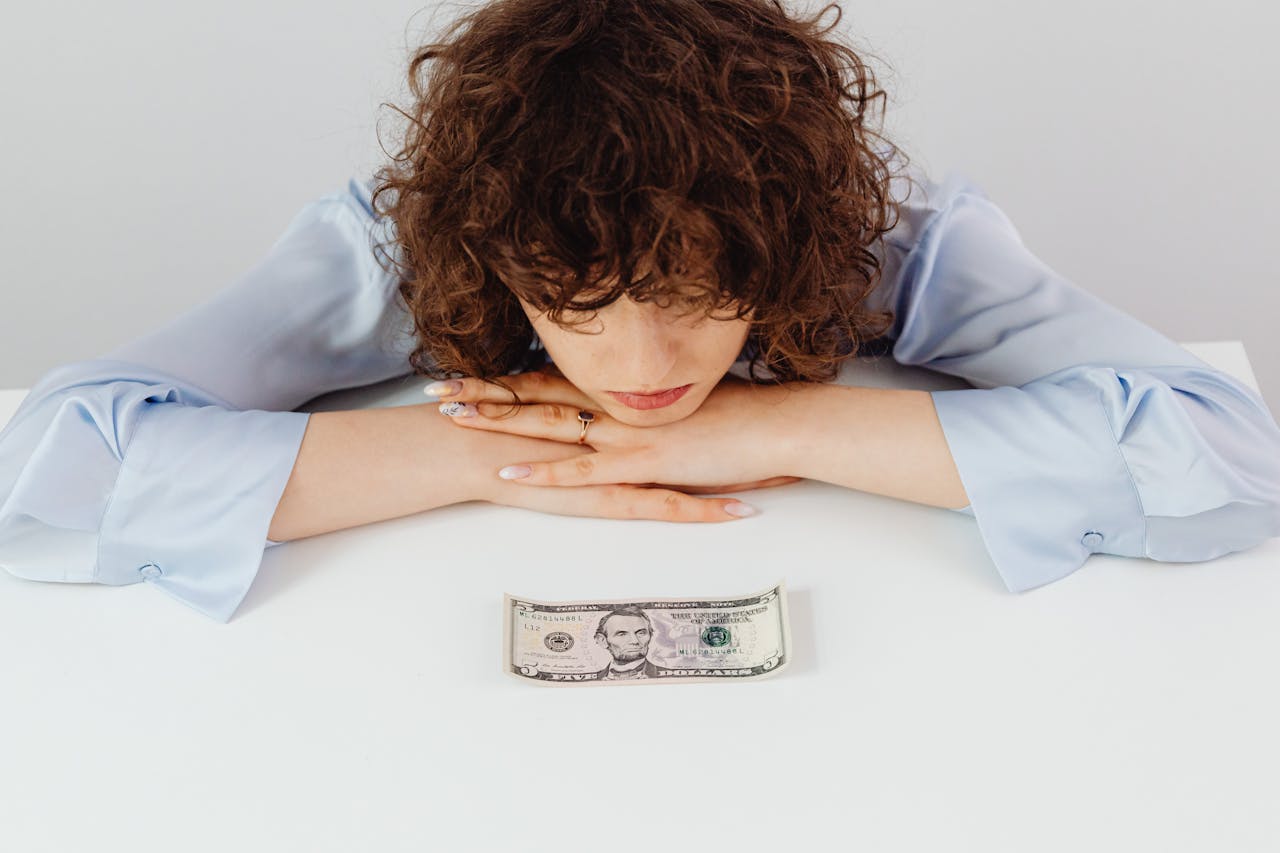 contact-img Caucasian woman with curly hair pondering over a five-dollar bill, expressing financial worry.